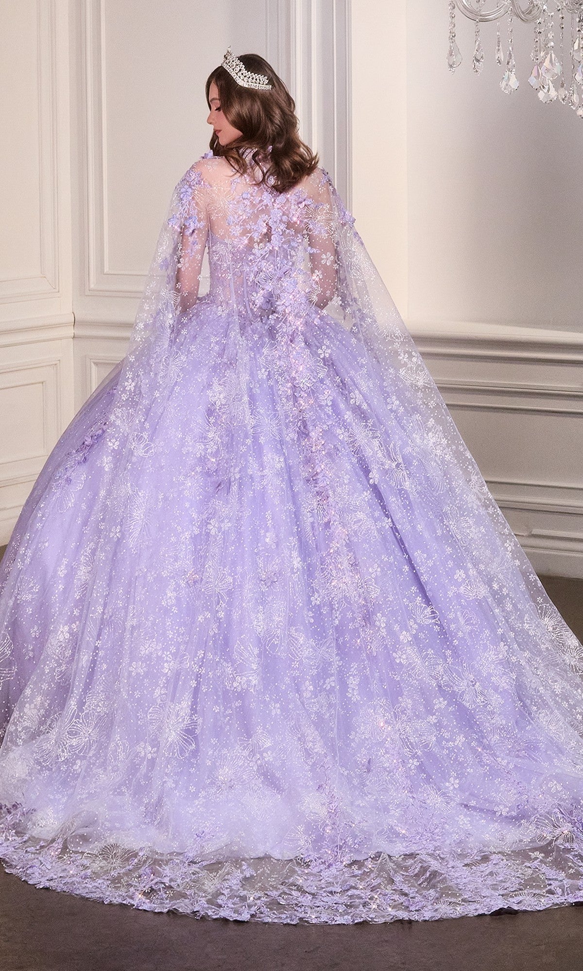 A woman in the Quinceanera Dress 15719 by Ladivine—a strapless ball gown with an ornate lace train—stands facing away in a white-paneled room, her silver tiara completing the elegant look.